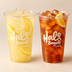 Two clear plastic cups from Halo Bagels Cafe filled with iced drinks, one with a yellow lemonade-like drink and lemon slices, and one with an iced coffee or cola in amber-brown liquid and a lemon wedge. Both cups are shown on a warm beige background.