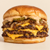 A close-up shot of a gourmet cheeseburger with two beef patties, melted cheddar cheese, caramelized onions, pickles, and a creamy sauce on a toasted bun, set against a solid light beige background.
