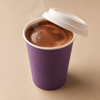 A purple paper coffee cup with a white lid, filled with steaming coffee, set against a solid beige background.